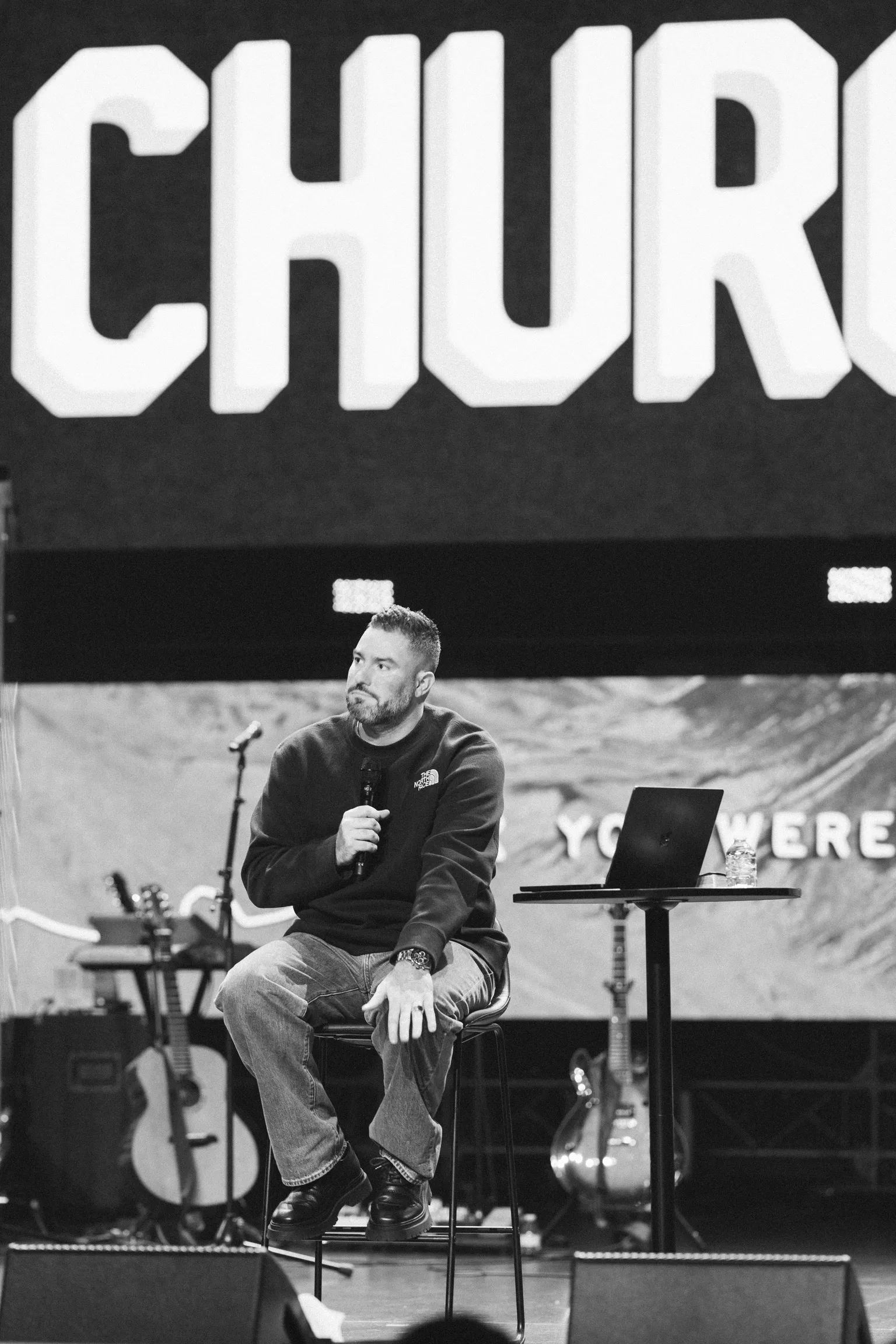 Black and white photo of a man seated on a stool speaking into a microphone, large letters spelling part of "CHURCH" visible on the screen behind him.