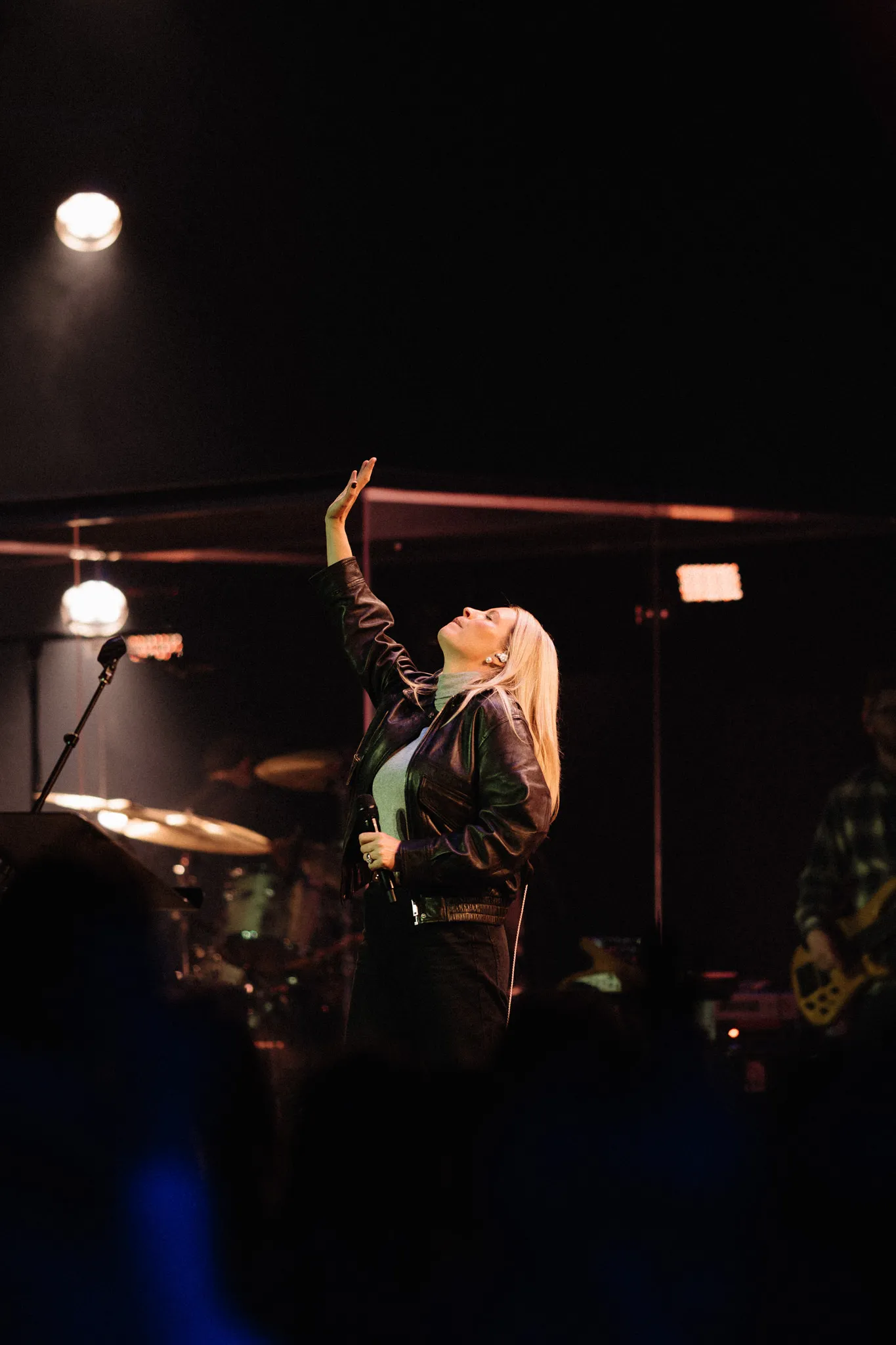 A woman on stage reaches one hand up during a worship moment under warm stage lighting.