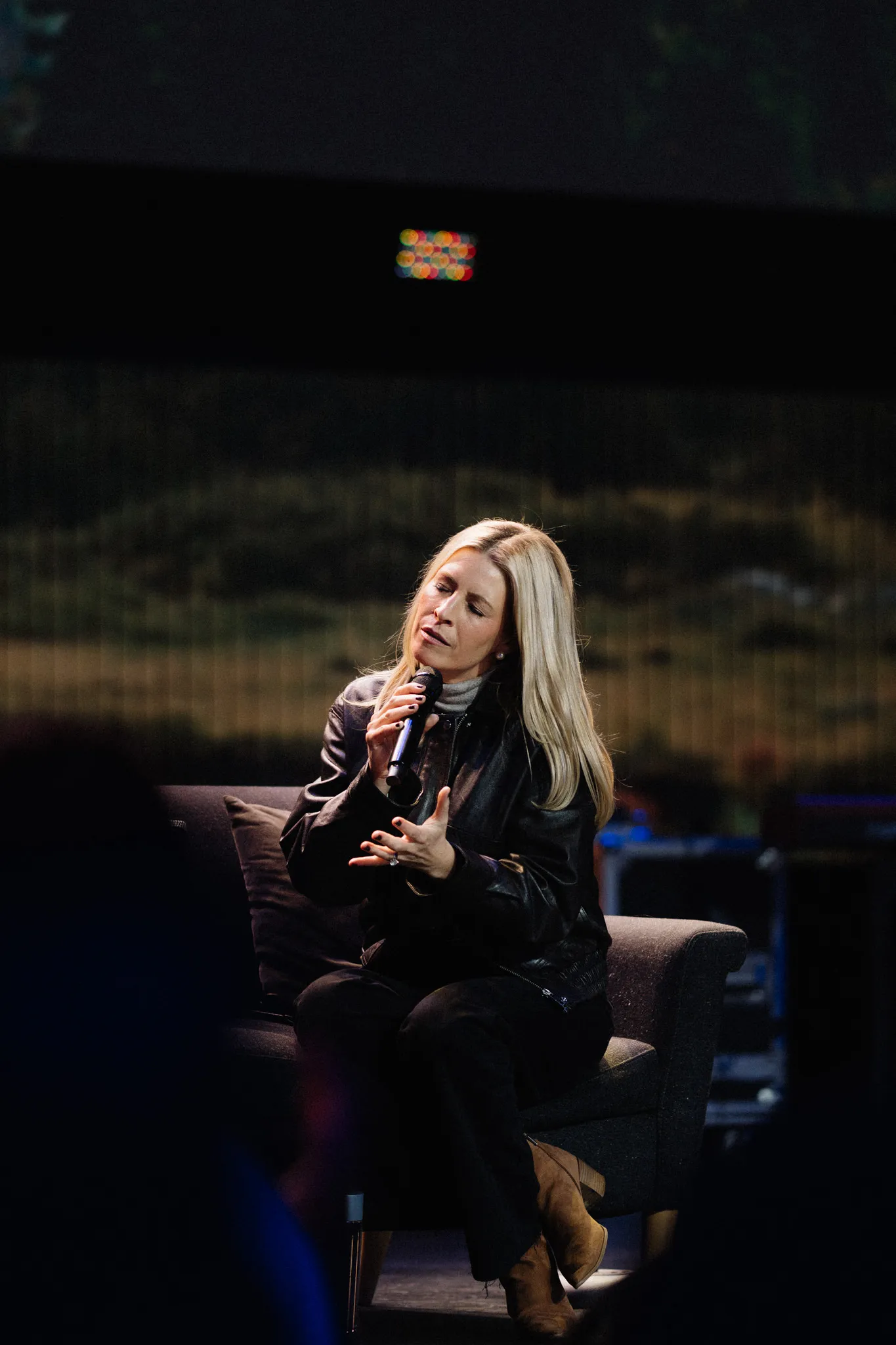 A woman seated on a couch on stage speaks into a microphone mid-gesture.