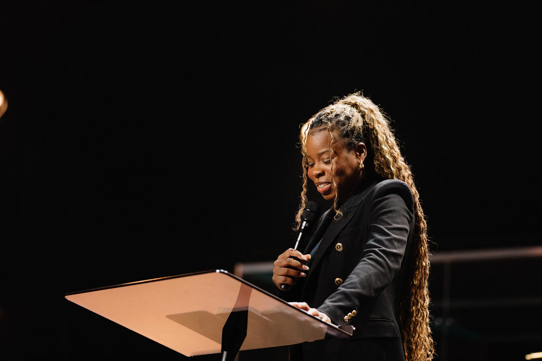 A woman with long braided hair speaks at a podium under warm stage lighting.