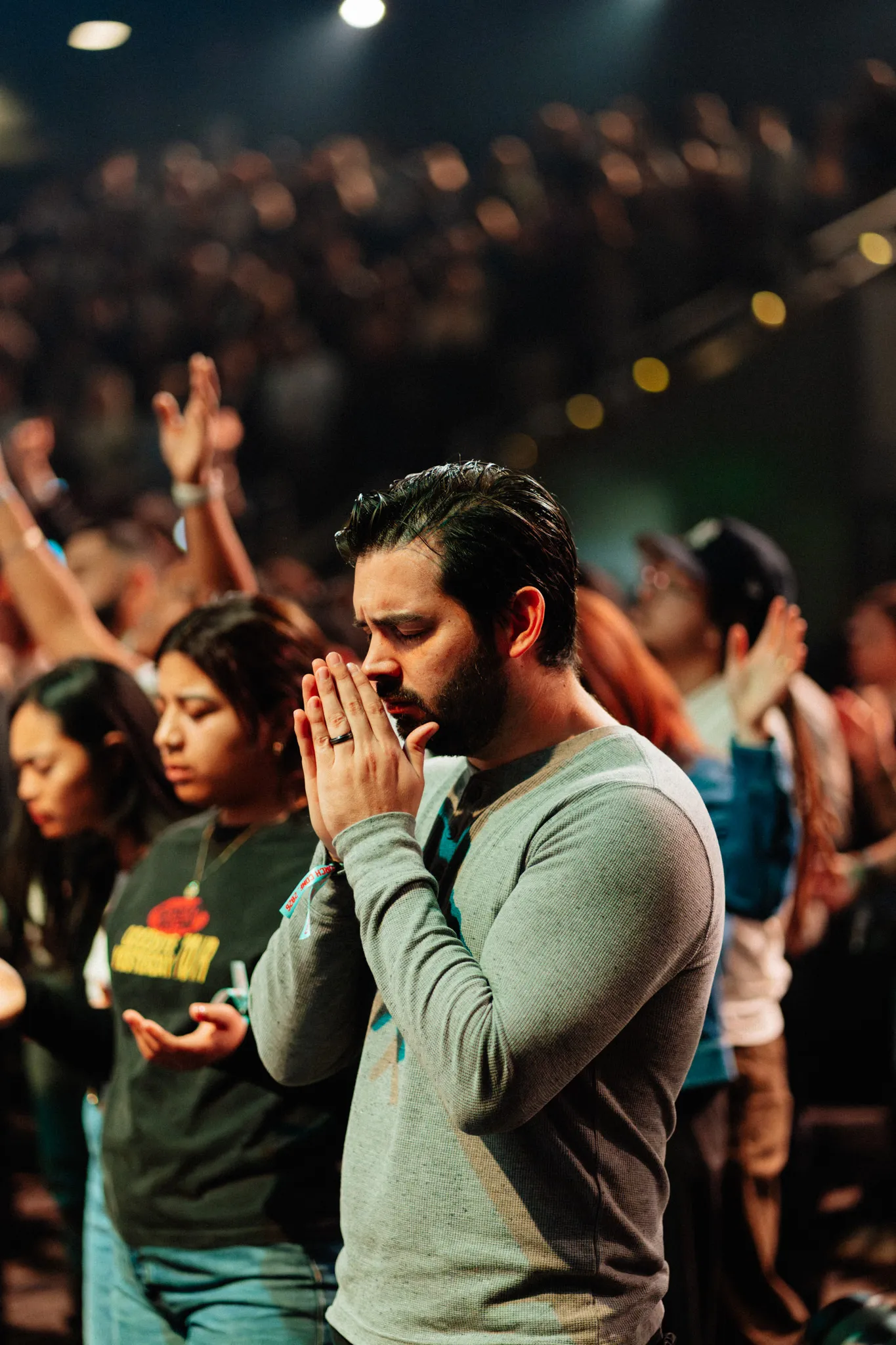 A man stands with his hands clasped in prayer, eyes closed, surrounded by a crowd with hands raised.