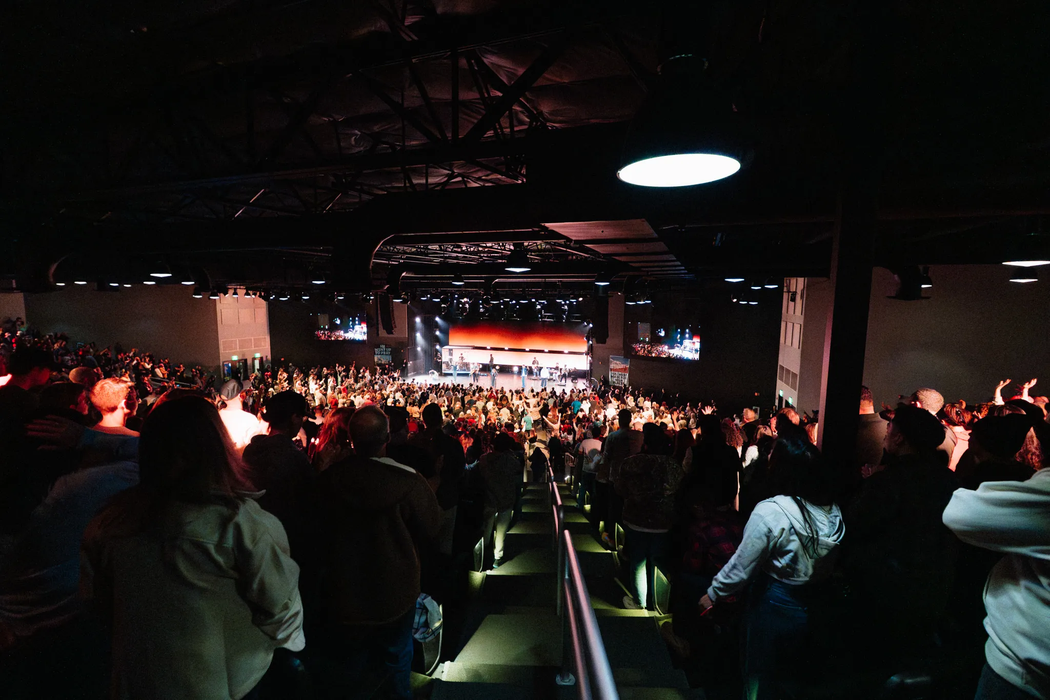 A wide view of a crowd inside a venue facing a lit stage, with people walking down stairs in the foreground.