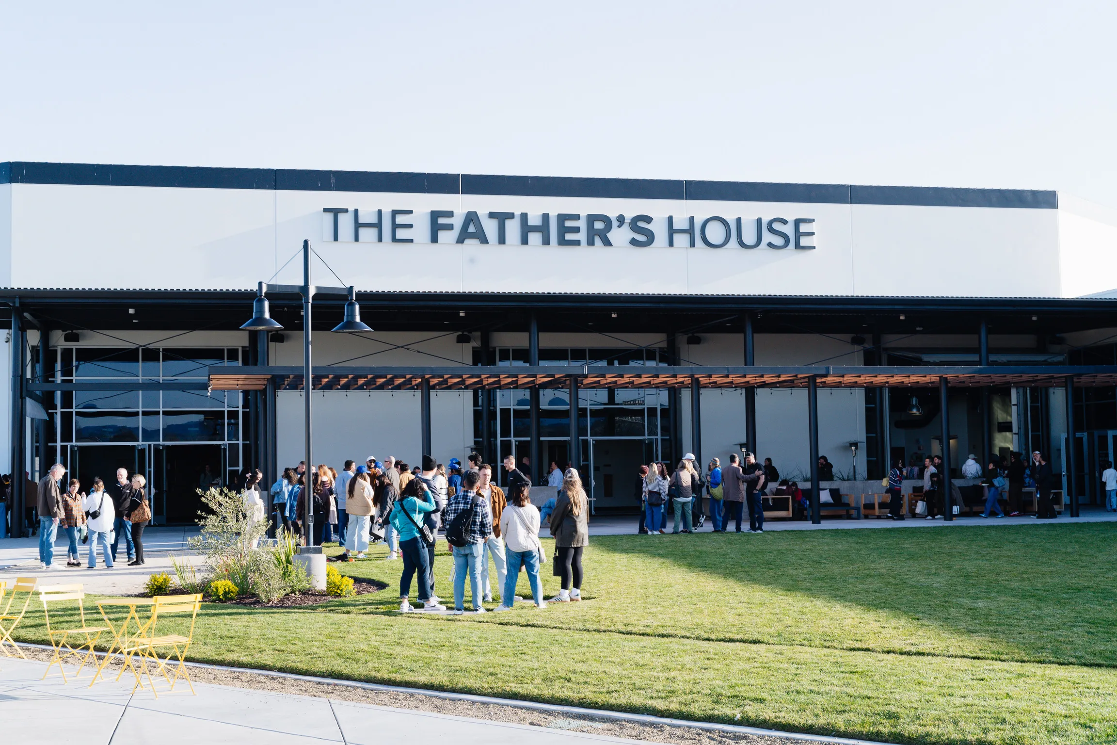 The exterior of The Father's House building on a sunny day, with people gathered on the lawn and patio.