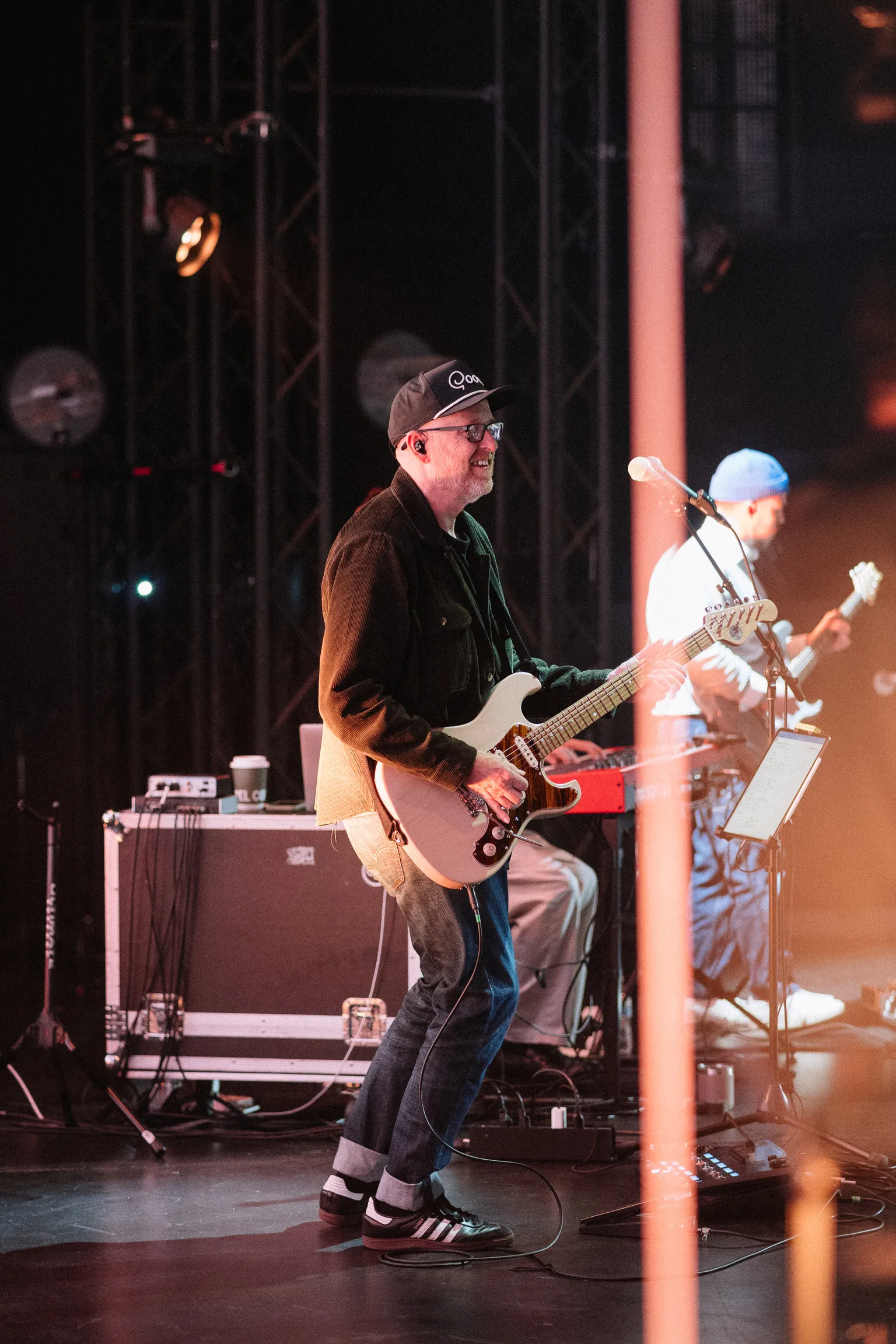A guitarist performs on stage under warm lighting with another musician behind him.