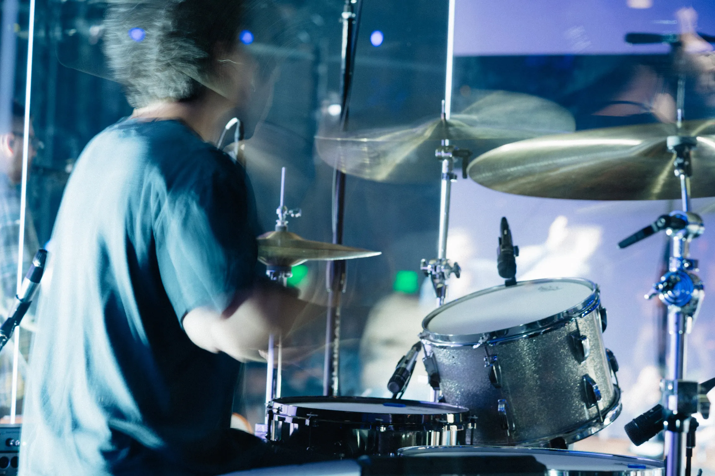 A motion-blurred photo of a drummer performing behind an acrylic sound booth.
