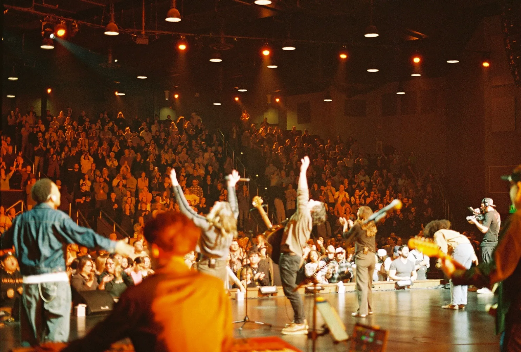 A wide color film photo of worship leaders on stage with arms raised, facing a crowd with hands up.