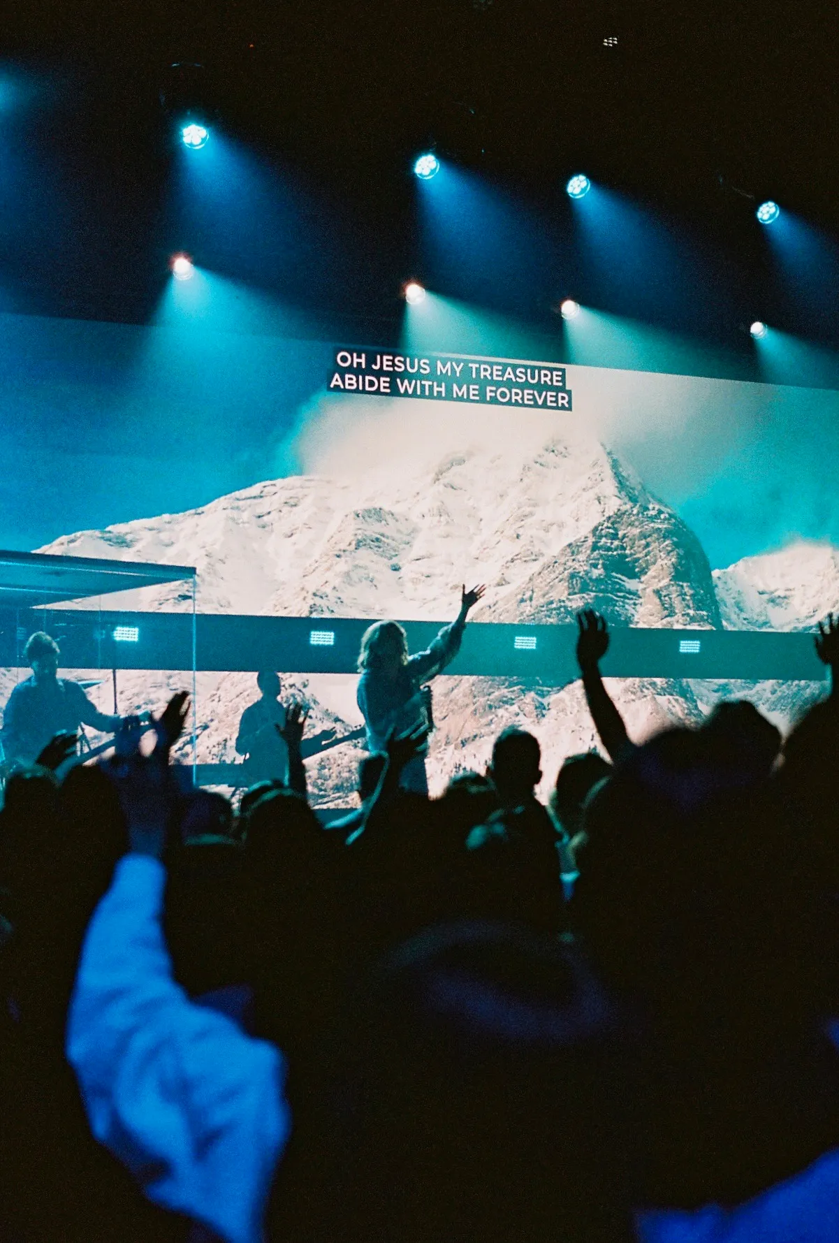 A crowd raises their hands toward a screen reading "Oh Jesus my treasure, abide with me forever" over an image of mountains.