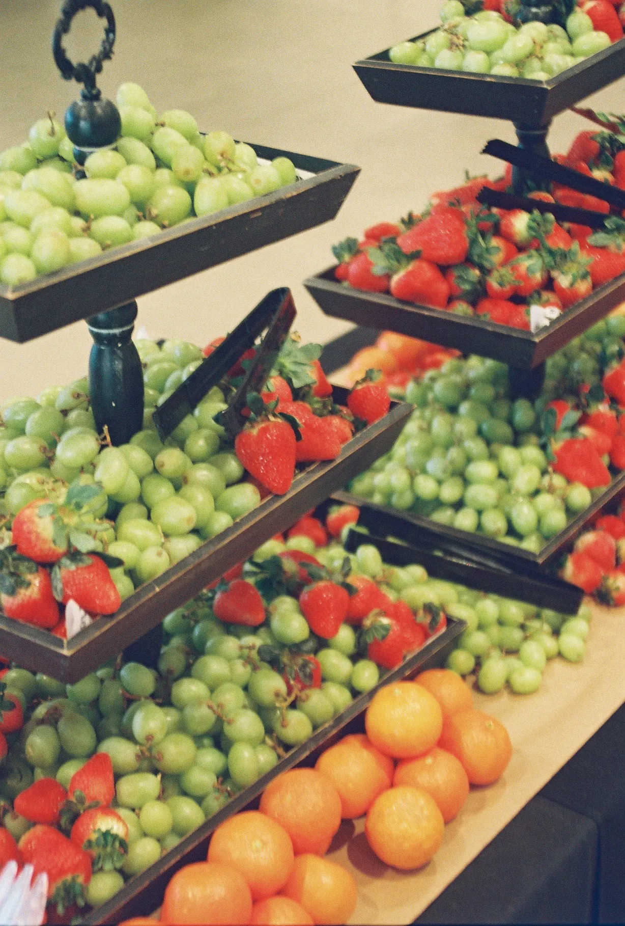 A film photo of tiered trays of grapes, strawberries, and oranges at a hospitality table.